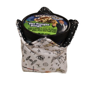 May include: A white fabric ice cream container holder with a black trim and a Star Wars themed print. The print features spaceships, stars, and the text "Meet the Galaxy". The holder is holding a pint of Ben & Jerry's ice cream called "The Tonight Dough".