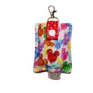 May include: A white fabric hand sanitizer holder with a red polka dot strap and a silver keychain. The holder features a colorful print of Mickey Mouse balloons.
