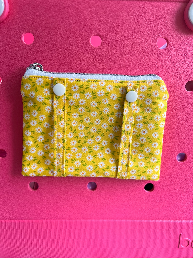 Pu&ograve; includere: Una piccola pochette gialla con cerniera con stampa di margherite bianche. La pochette ha due bottoni a pressione bianchi sul davanti.