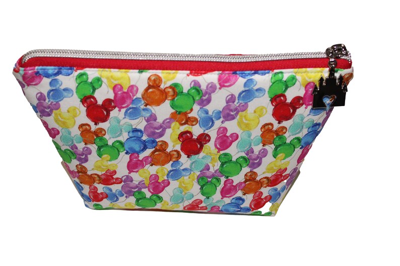 May include: A white zippered pouch with a red zipper and a black castle charm. The pouch is covered in a pattern of colorful Mickey Mouse balloons.