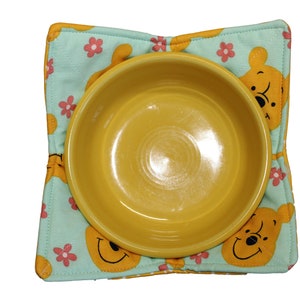 May include: A yellow ceramic bowl sits on a turquoise blue fabric placemat with a repeating pattern of cartoon bears and pink flowers.