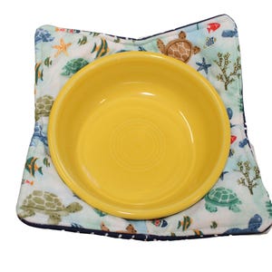 May include: A yellow ceramic bowl with a white and blue patterned fabric placemat featuring sea turtles, fish, and starfish.