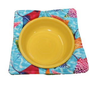 Può includere: Una ciotola di ceramica gialla su un sottopentola di stoffa blu e rosa con un motivo di razze colorate.