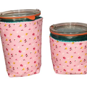 May include: Two glass jars with clear lids and orange silicone seals, each encased in a pink fabric sleeve featuring a small floral pattern with red and yellow blossoms. The fabric sleeves have a teal interior lining visible at the top rim.