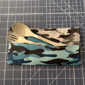 May include: A blue and black camouflage fabric pouch with a zipper closure and two black buttons. The pouch contains a silver fork and a white plastic spoon.