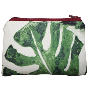 May include: A rectangular white pouch with a large green leaf pattern. The zipper is a dark red color. The pouch is likely made of fabric and is designed for carrying small items.