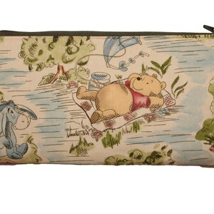 May include: Rectangular zippered pouch featuring a whimsical scene with Winnie the Pooh and Eeyore. The fabric has a light beige background with a colorful illustration of a picnic setting with trees, a kite, and a river. The zipper is dark green.
