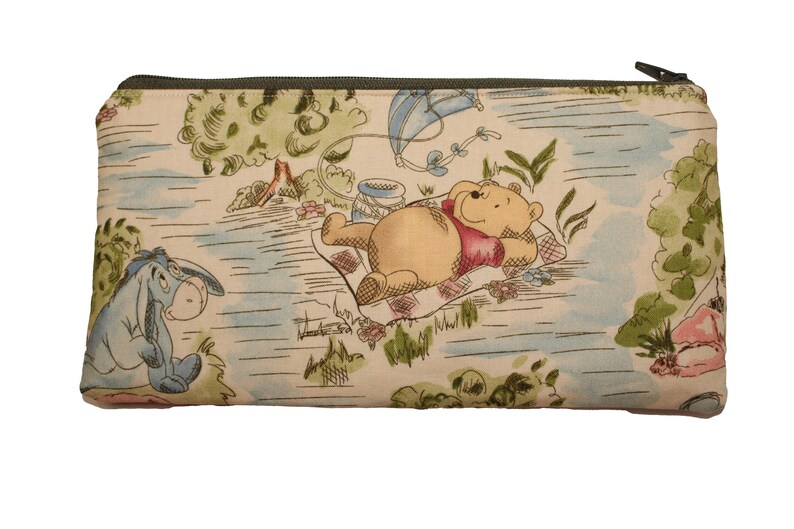May include: Rectangular zippered pouch featuring a whimsical scene with Winnie the Pooh and Eeyore. The fabric has a light beige background with a colorful illustration of a picnic setting with trees, a kite, and a river. The zipper is dark green.