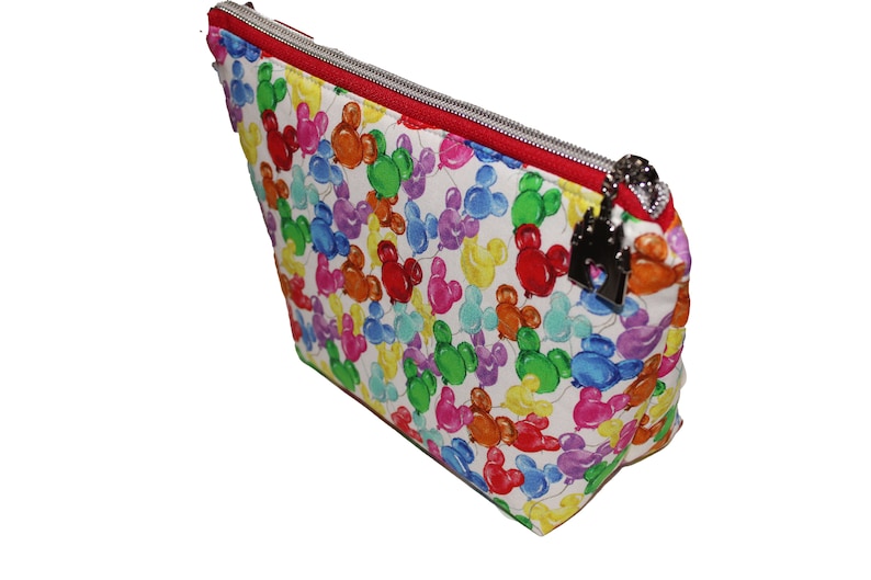 May include: A white zippered pouch with a red zipper and a silver charm. The pouch is covered in a colorful pattern of Mickey Mouse heads shaped like balloons.