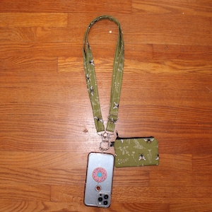 Puede incluir: Una correa de teléfono verde con un patrón de abejas y flores, unida a una funda de teléfono transparente con una pegatina de donut y una pequeña bolsa verde con cremallera con el mismo patrón de abejas y flores.