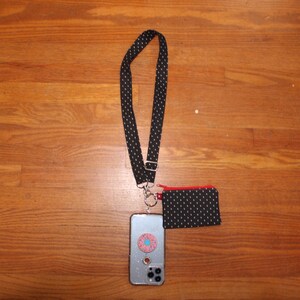 May include: A black and white polka dot phone strap with a matching small pouch. The strap is attached to a silver phone case with a pink and blue donut sticker.
