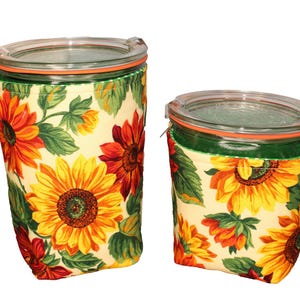 May include: Two glass jars with clear lids, each encased in a fabric sleeve featuring a sunflower pattern. The fabric displays yellow and orange sunflowers with green leaves on a cream background. The jars are of different heights and widths.