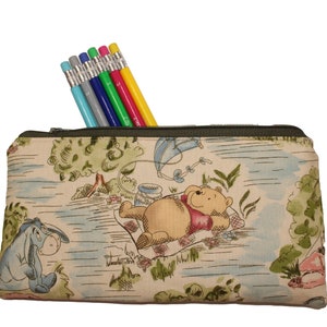 May include: A rectangular pencil case with a green zipper, filled with six pencils. The case features a Winnie the Pooh design with Eeyore, Pooh Bear, and a kite. The pencils are in various colors, including gray, blue, green, red, and yellow.