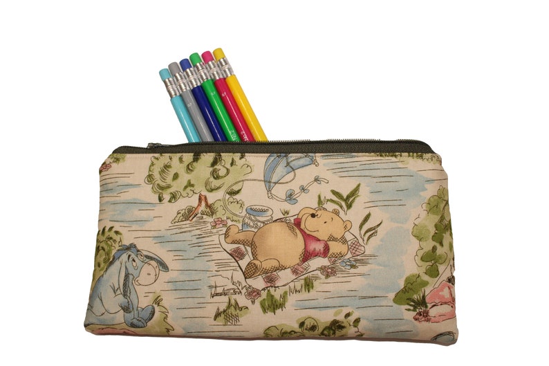 May include: A rectangular pencil case with a green zipper, filled with six pencils. The case features a Winnie the Pooh design with Eeyore, Pooh Bear, and a kite. The pencils are in various colors, including gray, blue, green, red, and yellow.