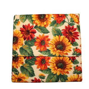 May include: A folded rectangular fabric item with a repeating pattern of sunflowers in shades of yellow, orange, and red, set against a cream-colored background. The sunflowers are interspersed with green leaves.