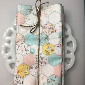 以下が含まれることがあります： ピンク、イエロー、ブルーの六角形のパッチワークデザインが施された、ファブリックナプキン2枚セット。ナプキンは紐で結ばれており、花柄が施されています。