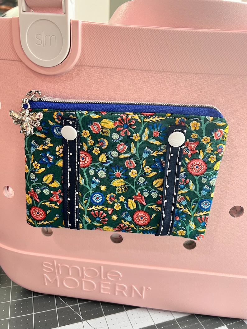 May include: A green and floral zippered pouch with blue trim and white buttons. The pouch is attached to a pink tote bag with the text "simple MODERN" on the bottom.