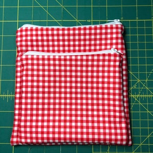 May include: Two red and white gingham fabric zippered pouches. The pouches are stacked on top of each other. The pouches are on a green cutting mat.