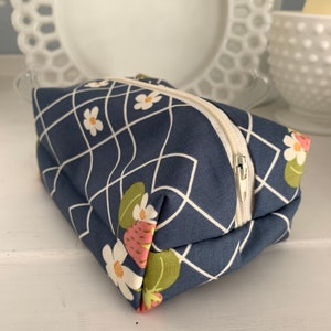 Puede incluir: Una pequeña bolsa azul con cremallera blanca. La bolsa tiene un patrón geométrico con líneas blancas y un diseño floral con flores blancas y fresas rosas.