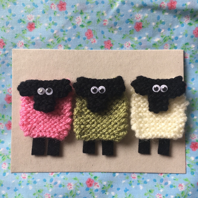 May include: Three knitted sheep with black faces and googly eyes. The sheep are pink, green, and white.