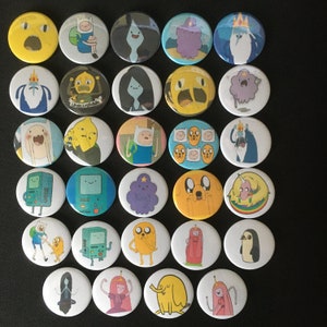 Op de afbeelding: Een set van 25 ronde, kleurrijke, bedrukte knoppen met personages uit de animatieserie Adventure Time. De knoppen zijn ongeveer 2,5 cm in diameter en bevatten verschillende personages, waaronder Finn, Jake, Prinses Bubblegum, IJskoning en Marceline.