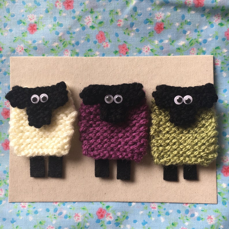 May include: Three knitted sheep ornaments, each with black faces and white eyes. The sheep are different colors: white, purple, and green.