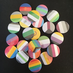 Op de afbeelding: Een verzameling ronde, kleurrijke speldjes met diverse ontwerpen. De speldjes tonen strepen, geometrische vormen en regenboogpatronen, die verschillende pride-vlaggen vertegenwoordigen. De speldjes zijn gerangschikt op een donkere ondergrond.