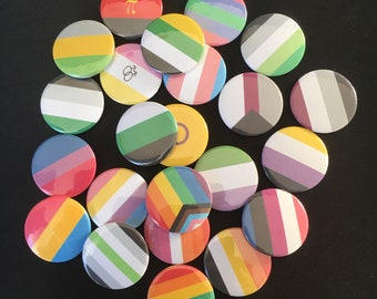 Puede incluir: Una colección de botones redondos y coloridos con varios diseños. Los botones presentan rayas, formas geométricas y patrones de arcoíris, que representan diferentes banderas del orgullo. Los botones están dispuestos sobre una superficie oscura.