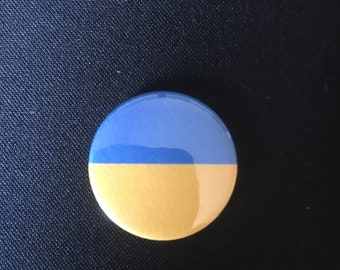 May include: A round button with a blue and yellow design. The top half of the button is blue and the bottom half is yellow.