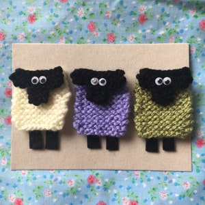 May include: Three knitted sheep ornaments with black faces and googly eyes. The sheep are white, purple, and green.
