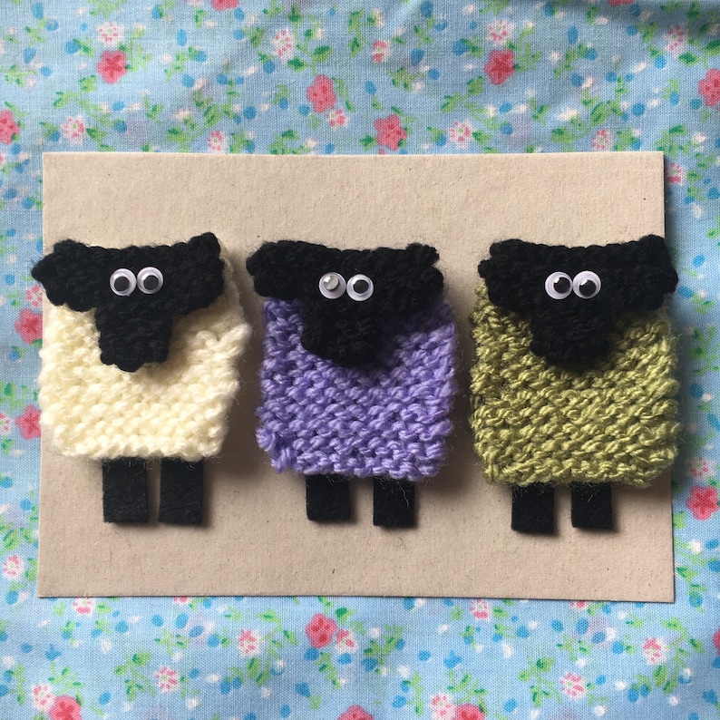 May include: Three knitted sheep ornaments with black faces and googly eyes. The sheep are white, purple, and green.