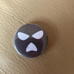 Può includere: Un bottone rotondo grigio scuro con un design a volto bianco stilizzato. Il volto presenta due occhi angolati e una bocca rivolta verso il basso. Il bottone è un design grafico semplice.