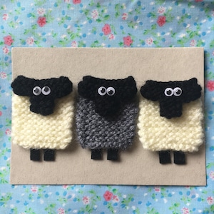 May include: Three knitted sheep, one white, one gray, and one white with a black face, are arranged in a row on a brown background. Each sheep has black eyes and a black nose.