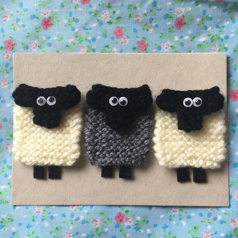 May include: Three knitted sheep, one white, one gray, and one white with a black face, are arranged in a row on a brown background. Each sheep has black eyes and a black nose.
