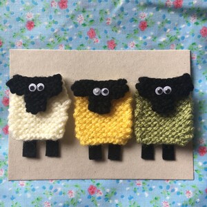 May include: Three knitted sheep with black faces and googly eyes. The sheep are white, yellow, and green.