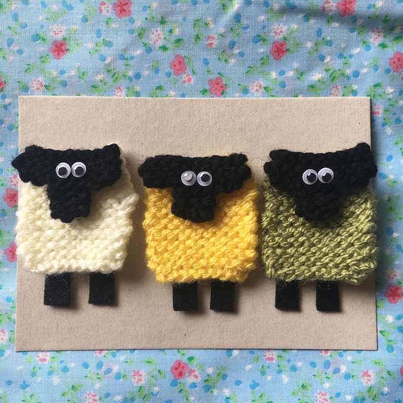 May include: Three knitted sheep with black faces and googly eyes. The sheep are white, yellow, and green.