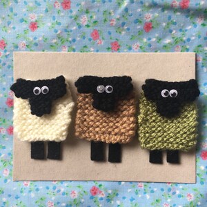 May include: Three knitted sheep ornaments, each with a black face and white eyes. The sheep are white, brown, and green.
