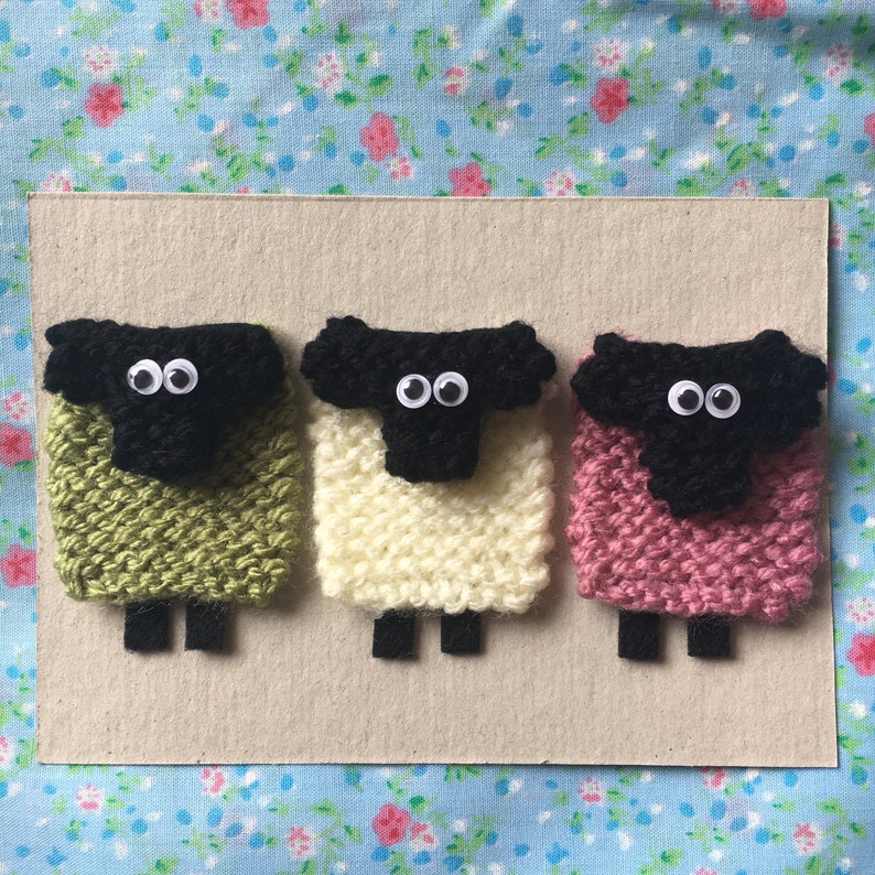 May include: Three knitted sheep with black faces and googly eyes. The sheep are green, white, and pink.