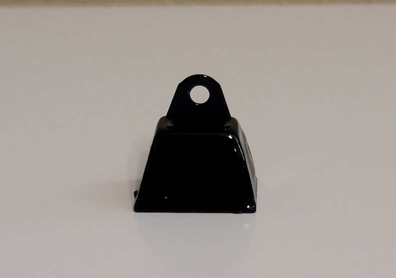 May include: A small, black metal bell with a square base and a loop at the top.