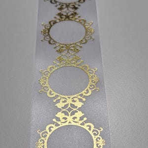 May include: White ribbon with a repeating pattern of gold filigree frames. The frames are oval-shaped with ornate detailing. The ribbon is likely used for crafts, gift wrapping, or embellishments.