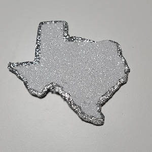 May include: A white glitter Texas state outline with a silver border.