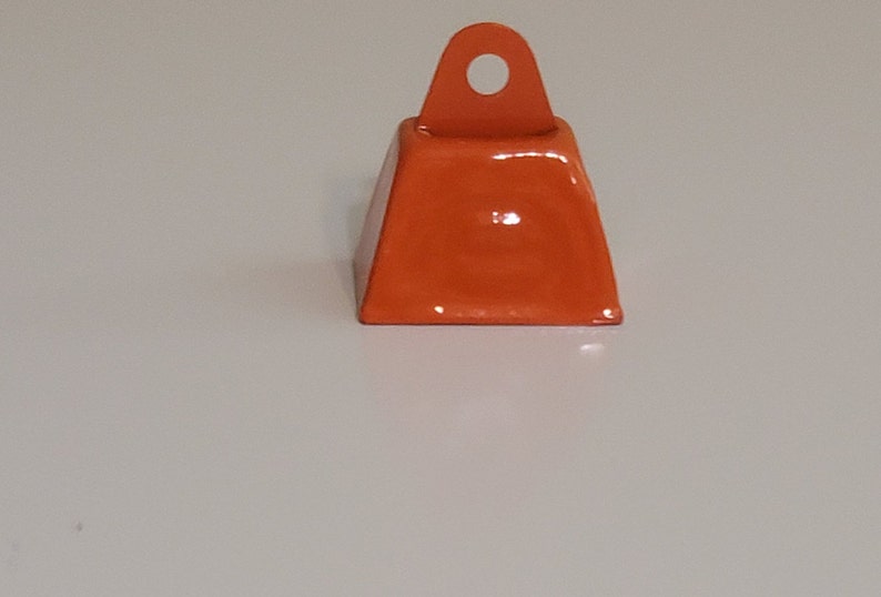 May include: A small, orange metal bell with a loop at the top. The bell is approximately 2.5 cm in diameter.
