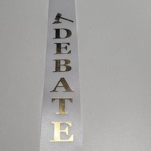 May include: White ribbon with gold lettering that spells out "DEBATE" vertically. A gold gavel icon is at the top. The ribbon is likely for an academic or competitive event.