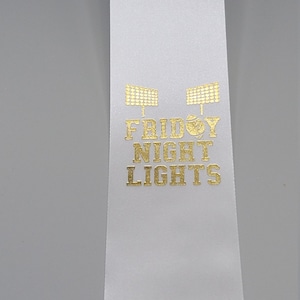 May include: White ribbon with gold lettering that reads "Friday Night Lights" and a graphic of a football. The graphic is repeated three times on the ribbon.