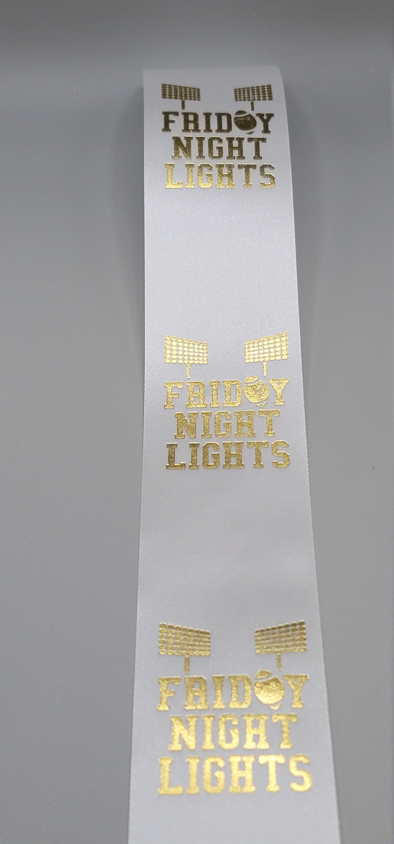 May include: White ribbon with gold lettering that reads "Friday Night Lights" and a graphic of a football. The graphic is repeated three times on the ribbon.