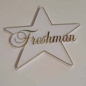 May include: White star-shaped sign with a gold outline and the word "Freshman" written in gold cursive lettering.