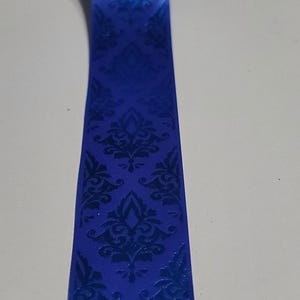 May include: A length of royal blue ribbon with a repeating dark blue damask pattern. The ribbon has a slight sheen, and the pattern is embossed, creating a textured effect. The ribbon is likely used for crafting or gift wrapping.