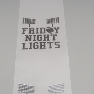 May include: White ribbon with the text "Friday Night Lights" printed in silver. The text is repeated three times on the ribbon. Each time the text is printed, there are two silver silhouettes of football stadium lights above the text and a silver football in the middle of the text.