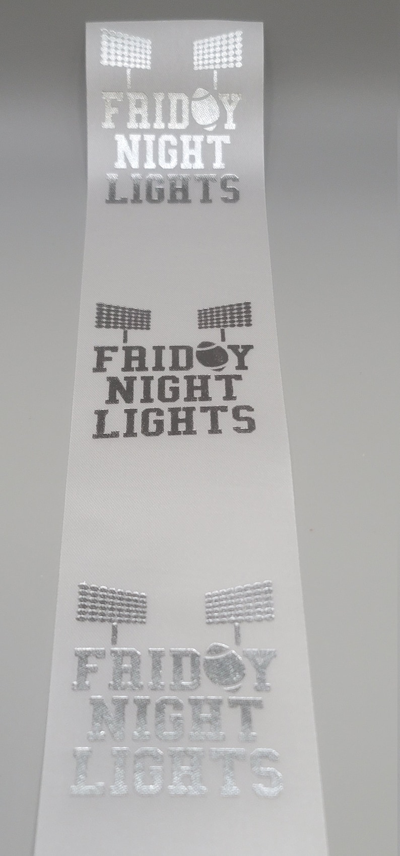 May include: White ribbon with the text "Friday Night Lights" printed in silver. The text is repeated three times on the ribbon. Each time the text is printed, there are two silver silhouettes of football stadium lights above the text and a silver football in the middle of the text.