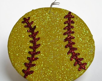 May include: A glittery yellow softball ornament with red stitching details. The ornament is round and has a string for hanging. The softball design is a festive decoration for sports fans.